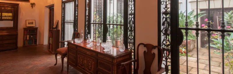 Interior de una sala de museo con un aparador de madera que exhibe cristalería, grandes ventanales con rejas de hierro que dan a un patio con plantas y mobiliario de época.