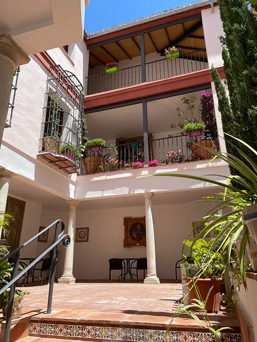 Patio interior del museo con paredes blancas, columnas clásicas, balcones con barandillas de hierro llenos de plantas en macetas y flores, suelo de baldosas de terracota y vigas de madera en el techo bajo un cielo azul
