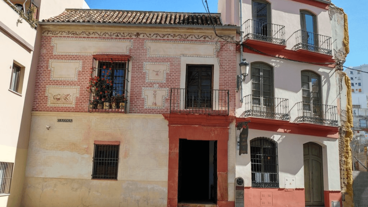 Fachada de edificio de dos plantas en color rosa y blanco con balcones de hierro, ventanas con rejas y una puerta central bajo un tejado de tejas.