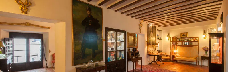 Sala de museo con techo de vigas de madera, un gran retrato en la pared, vitrinas de cristal y muebles de época bajo una iluminación cálida