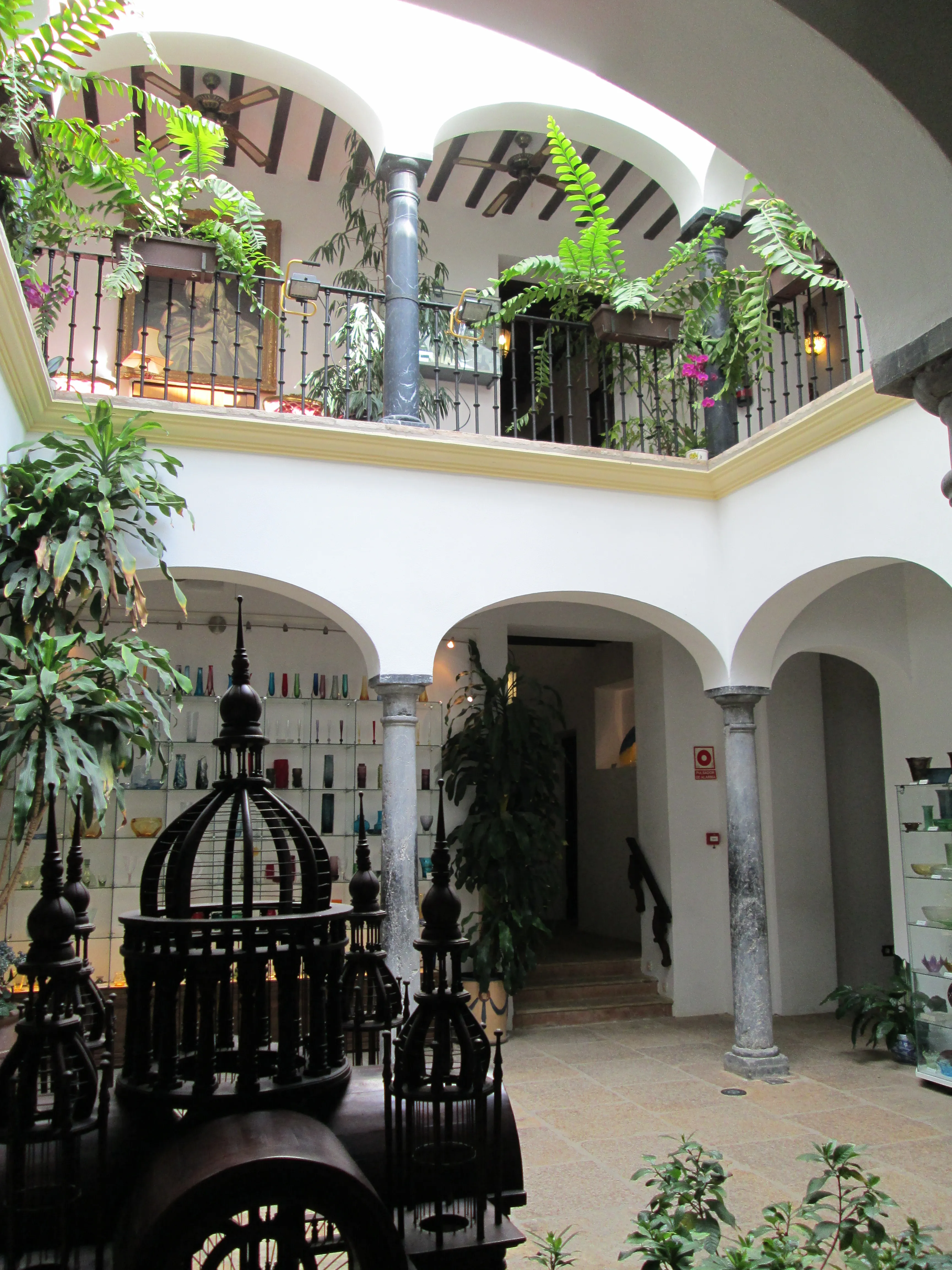 Patio interior de dos plantas con arcadas blancas, barandillas de hierro en el balcón superior, frondosas plantas verdes y una jaula de pájaros decorativa de hierro en primer plano, con vitrinas de cristal visibles a través de los arcos