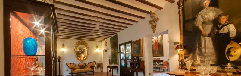 Vista interior de una estancia de época con vigas de madera en el techo, muebles antiguos, un espejo grande y un jarrón de vidrio azul en una vitrina.