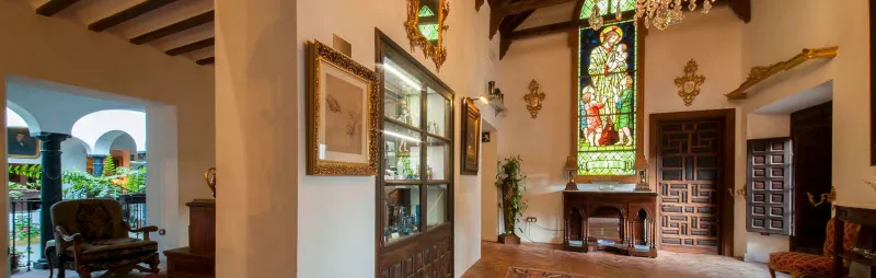 Interior de un museo con vigas de madera en el techo, un ventanal con vitrales de figuras coloridas, vitrinas de cristal, obras de arte enmarcadas y mobiliario de época en una casa histórica.