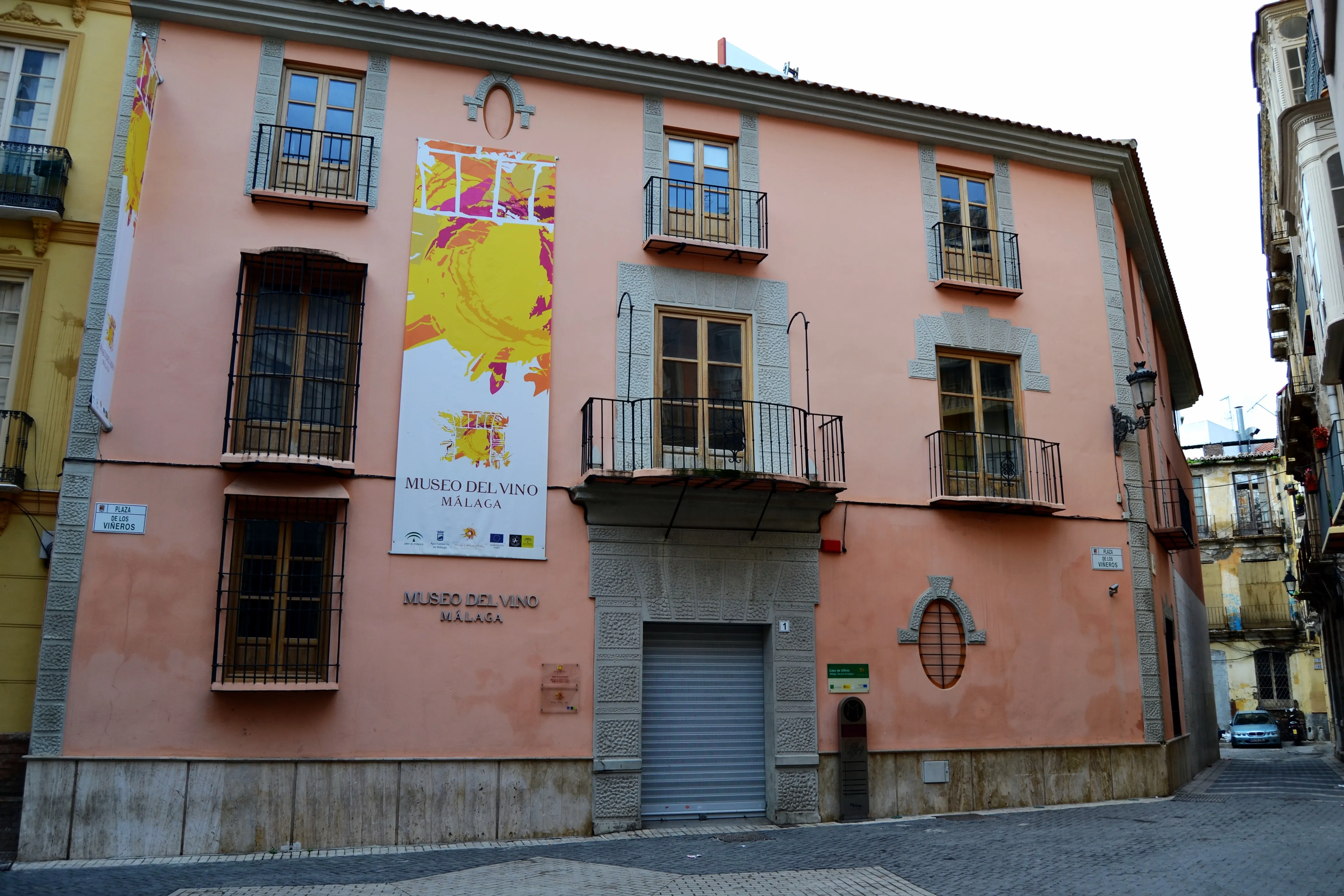 Fachada rosa del edificio del Museo del Vino Málaga con una gran pancarta promocional amarilla y blanca, balcones de hierro y una entrada a pie de calle en una calle estrecha.