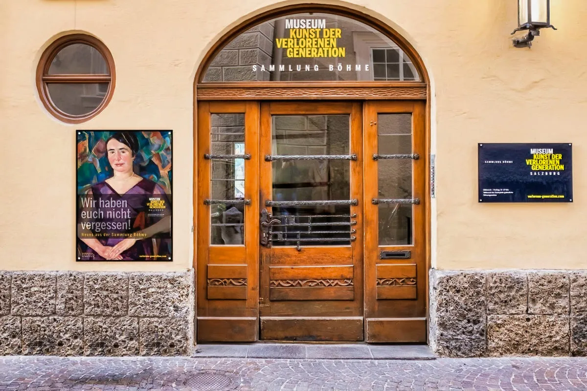 Entrada de doble puerta de madera del Museum Kunst der Verlorenen Generation con una ventana arqueada encima que muestra el nombre del museo, flanqueada por un cartel y un letrero en una pared beige.