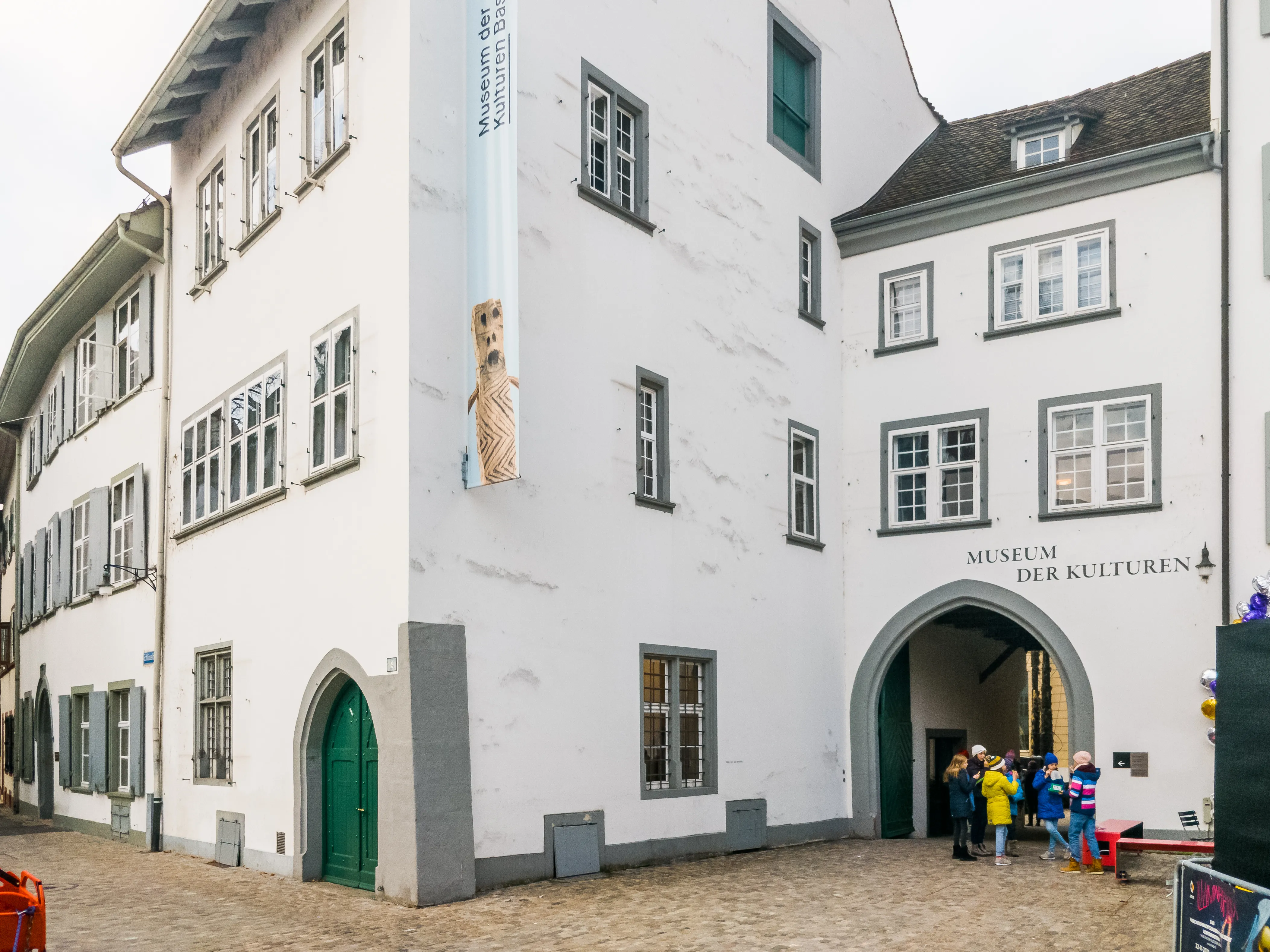 Fachada blanca con entrada en arco que muestra el letrero del Museum der Kulturen, con visitantes reunidos en la entrada y un cartel alto en la pared de la esquina