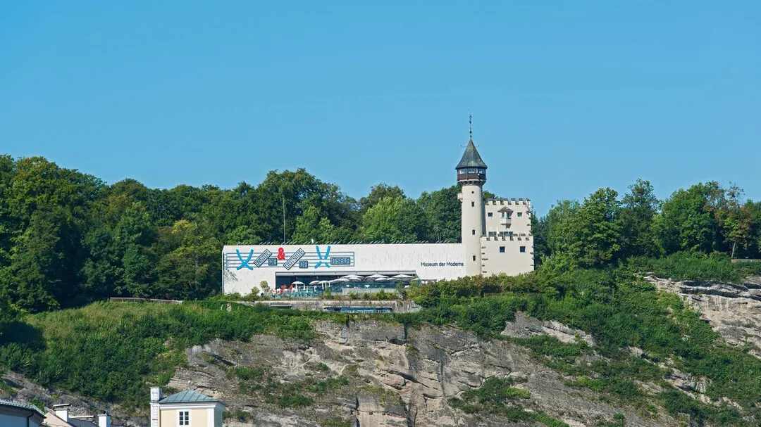 Breed buitenaanzicht van het gebouw van het Museum der Moderne Salzburg met een kleurrijke muurschildering op de witte gevel, bevestigd aan een historische stenen toren, gelegen op een rotsachtige, beboste heuvel onder een helderblauwe lucht.