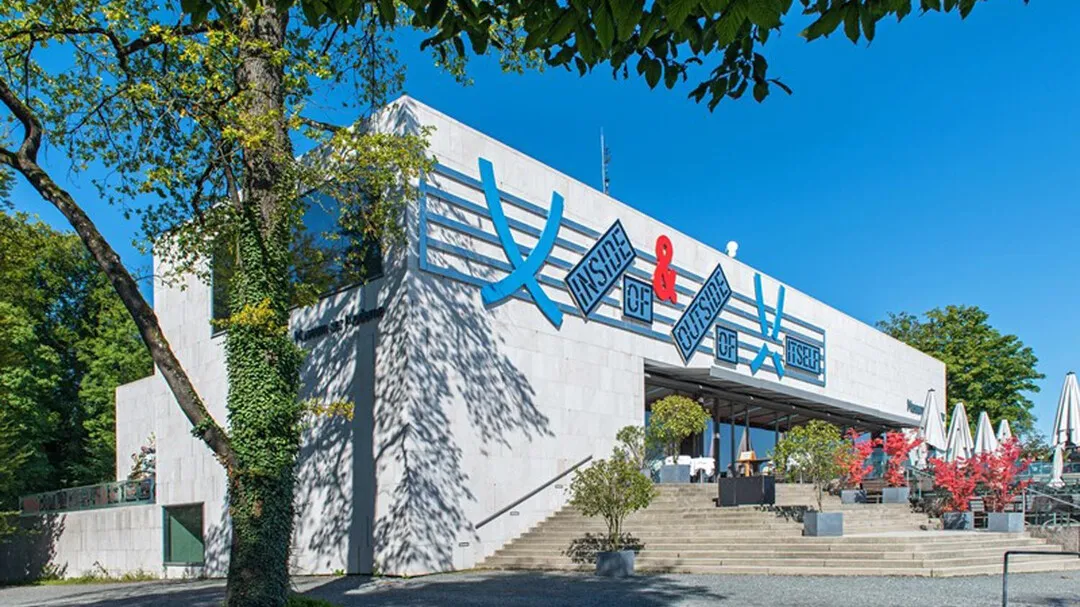 Witte modernistische gevel met een groot blauw geometrisch museumlogo, stenen trappen die naar de ingang leiden, bomen en een helderblauwe lucht