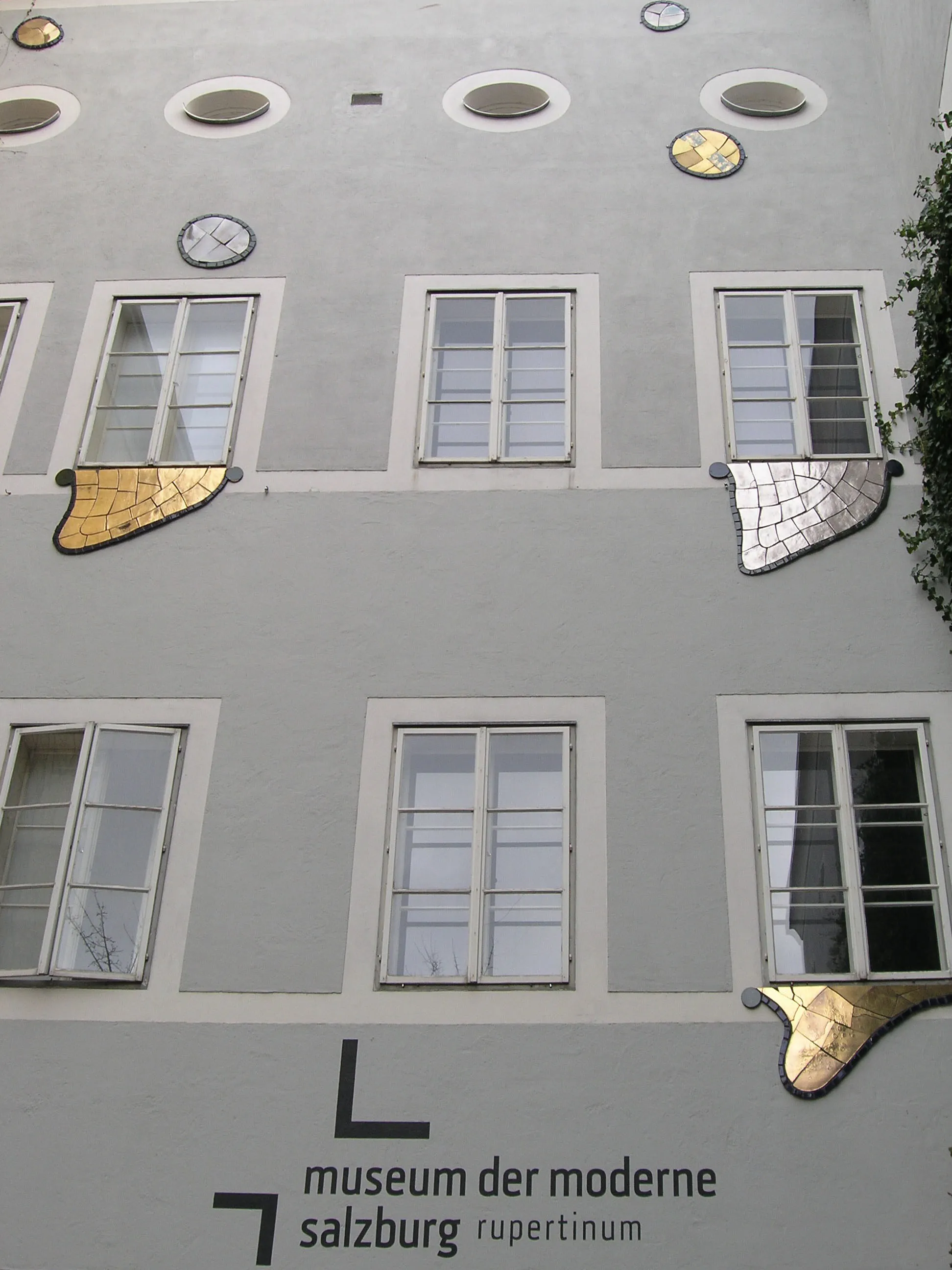 Jasnoszara fasada budynku z okrągłymi i prostokątnymi oknami, charakterystycznymi złotymi i srebrnymi mozaikami pod wybranymi oknami oraz oznaczeniem museum der moderne salzburg rupertinum na dole.