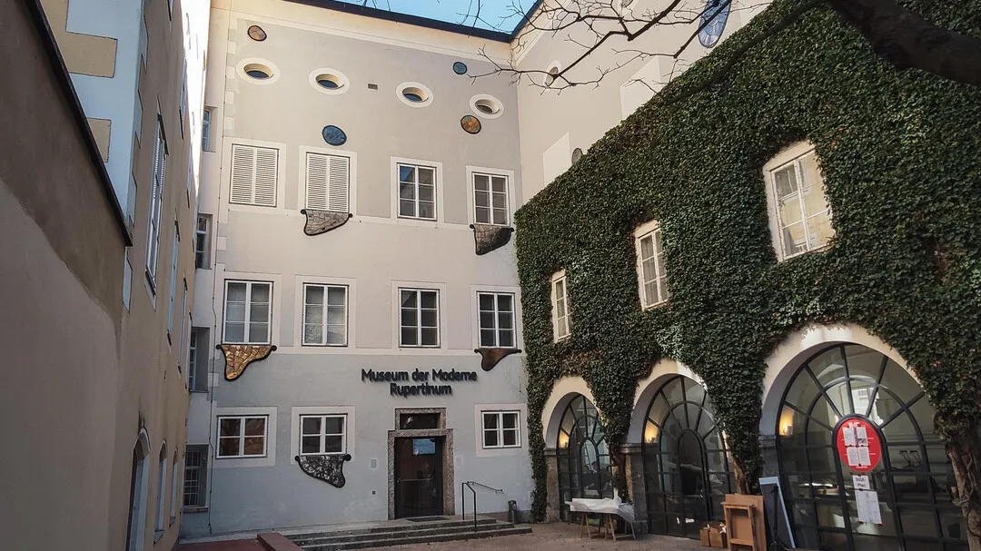 Widok z zewnątrz na Museum der Moderne Rupertinum przedstawiający białą fasadę z nazwą muzeum nad wejściem oraz przylegający budynek porośnięty bluszczem z łukowatymi drzwiami.