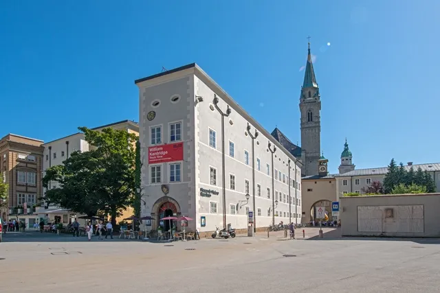 Widok z zewnątrz na Museum der Moderne Salzburg Rupertinum, biały barokowy budynek z czerwonym banerem, z widoczną w tle zieloną wieżą kościelną na tle bezchmurnego błękitnego nieba