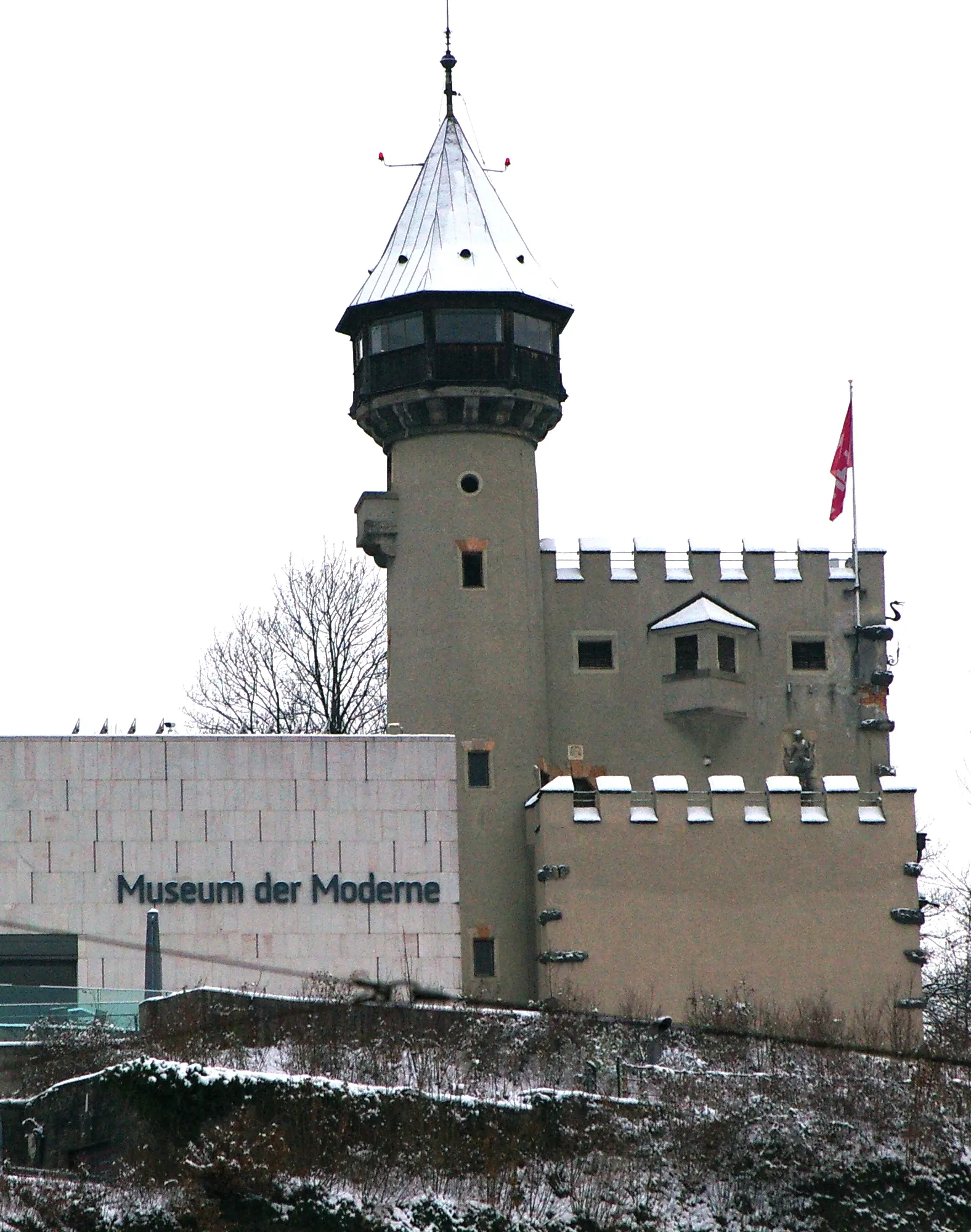 Widok z zewnątrz na Museum der Moderne Salzburg, przedstawiający zabytkową wieżę ze stożkowym dachem i blankami obok nowoczesnego białego budynku z szyldem muzeum, ze śniegiem na ziemi i dachach.