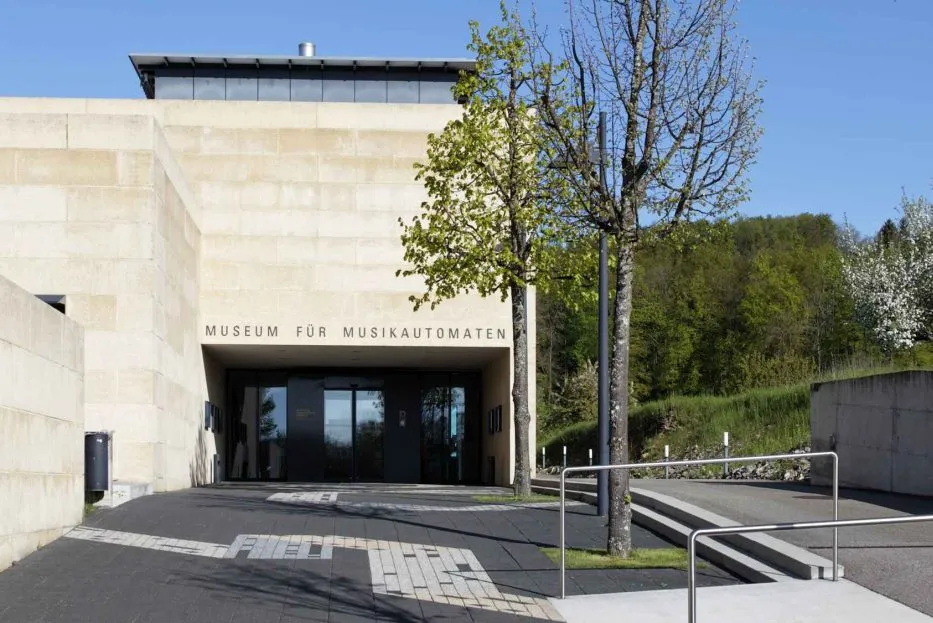 Beige stenen gevel van het Museum für Musikautomaten met de ingang en bewegwijzering zichtbaar onder een helderblauwe lucht.