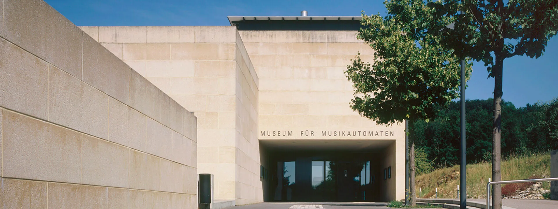 Moderne beige stenen gevel van een gebouw met de tekst 'Museum für Musikautomaten' boven een glazen entree, geflankeerd door groene bomen onder een blauwe lucht