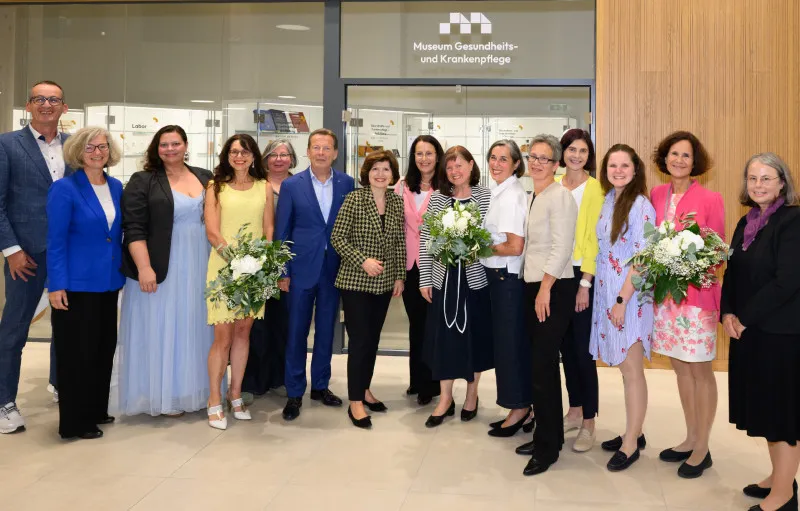 Group of people standing in front of a museum entrance with a sign reading 'Museum Gesundheits- und Krankenpflege' holding bouquets of flowers