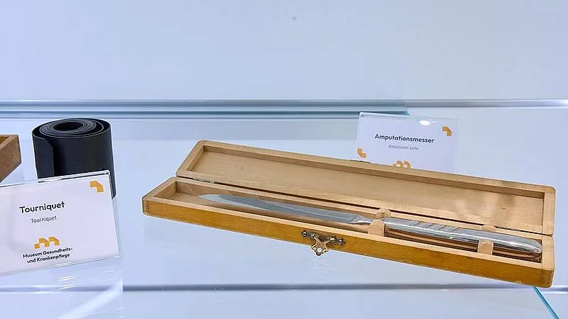 A wooden box containing a surgical instrument on display in a glass case with informational cards.