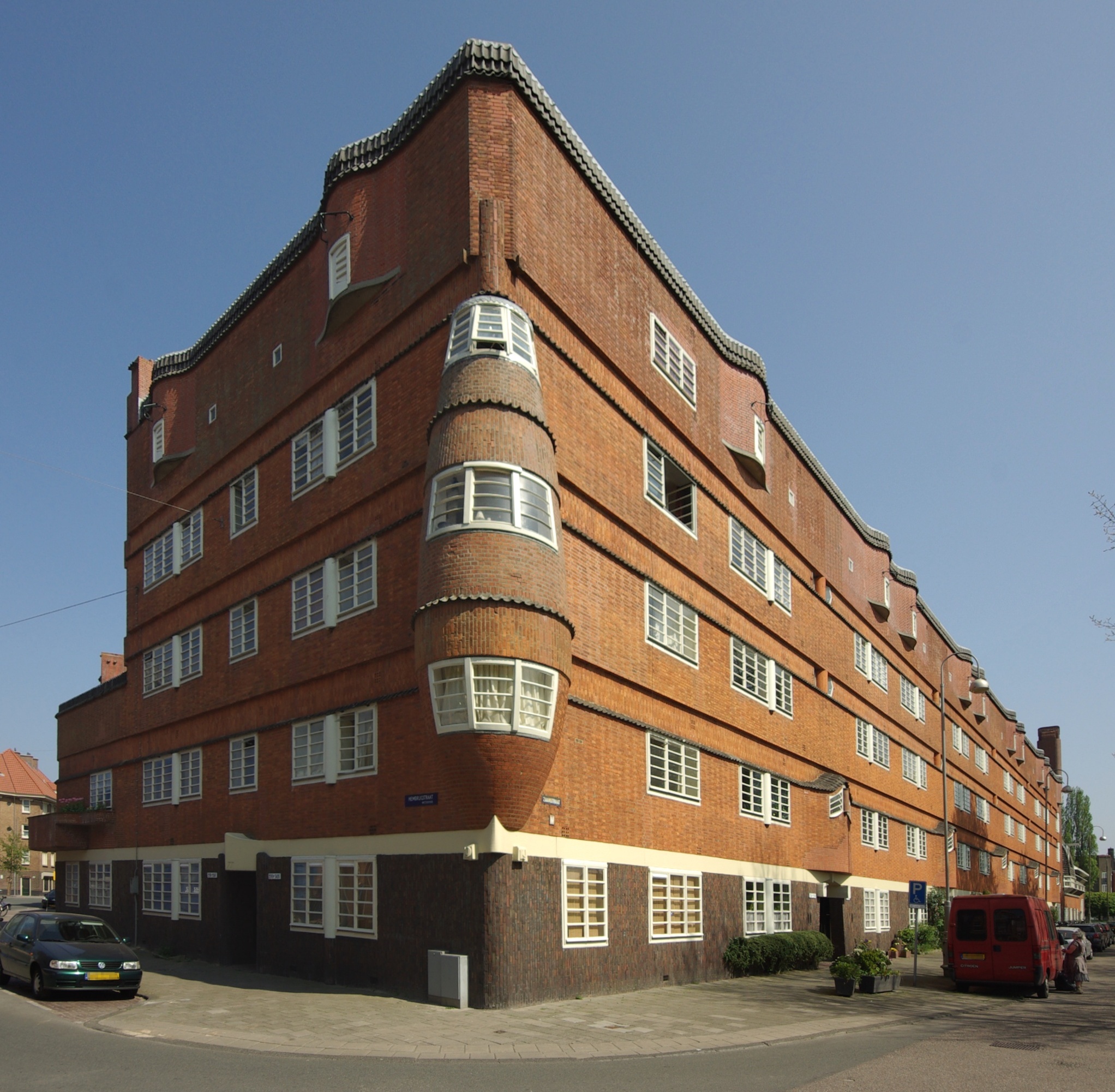 Red-brick Amsterdam School style building with rounded corner, white-framed windows, and street-level entrance under clear blue sky.