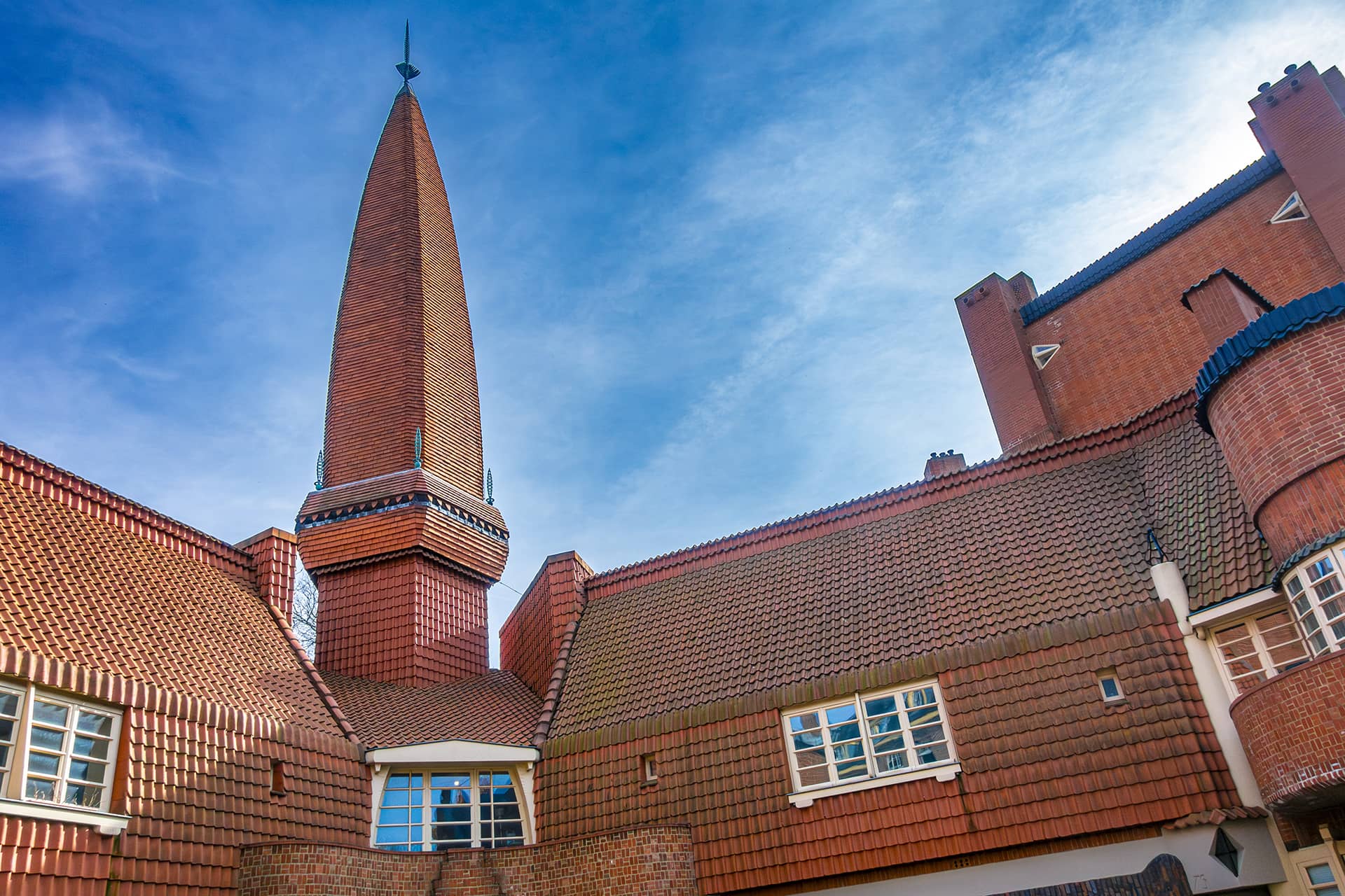 Red brick Amsterdam School-style facade of Museum Het Schip featuring a tall spire, tiled roof, and white-framed windows against a bright blue sky.