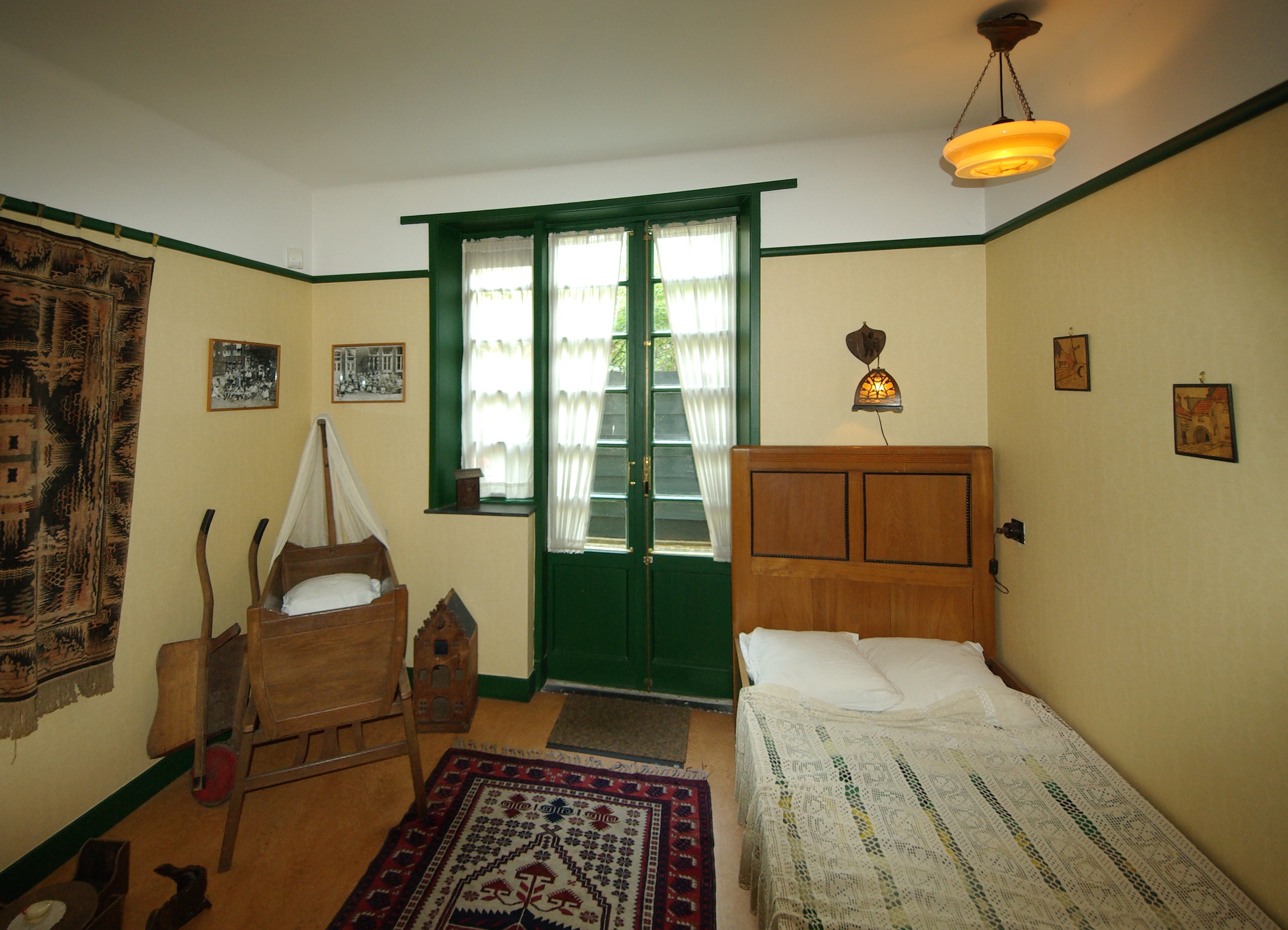 A bedroom with a wooden bed, a cradle, and green-trimmed windows, featuring period furnishings and a patterned rug.