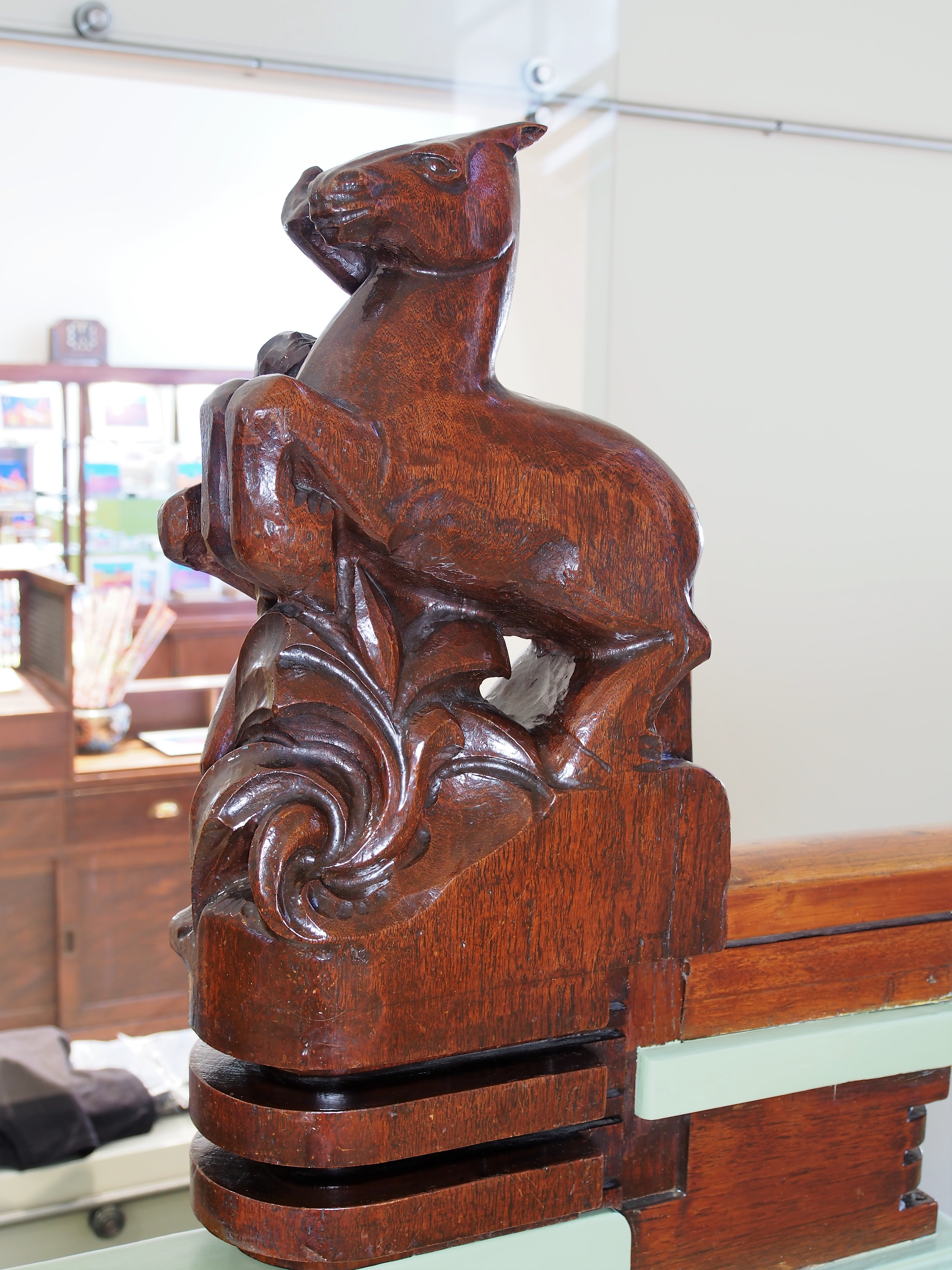Talla de madera de una figura de cabra con intrincados patrones florales en muebles, expuesta en el interior de un museo con armarios al fondo.