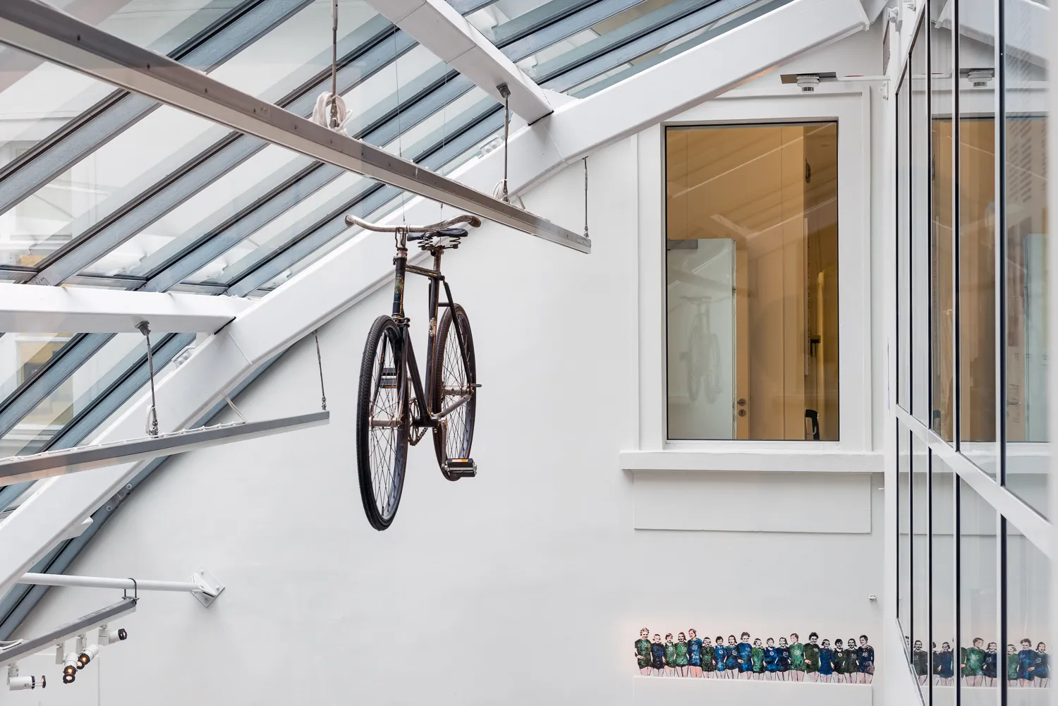 Een fiets die aan een glazen plafond hangt in een modern gebouw met witte muren en ramen.