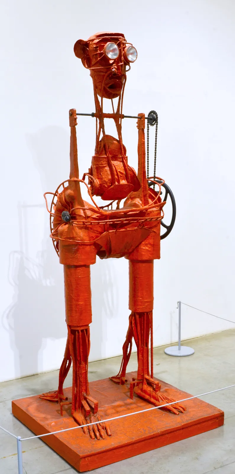 Una escultura alta, roja, de alambre y tela de una figura humanoide con partes mecánicas, expuesta sobre una plataforma en una galería.