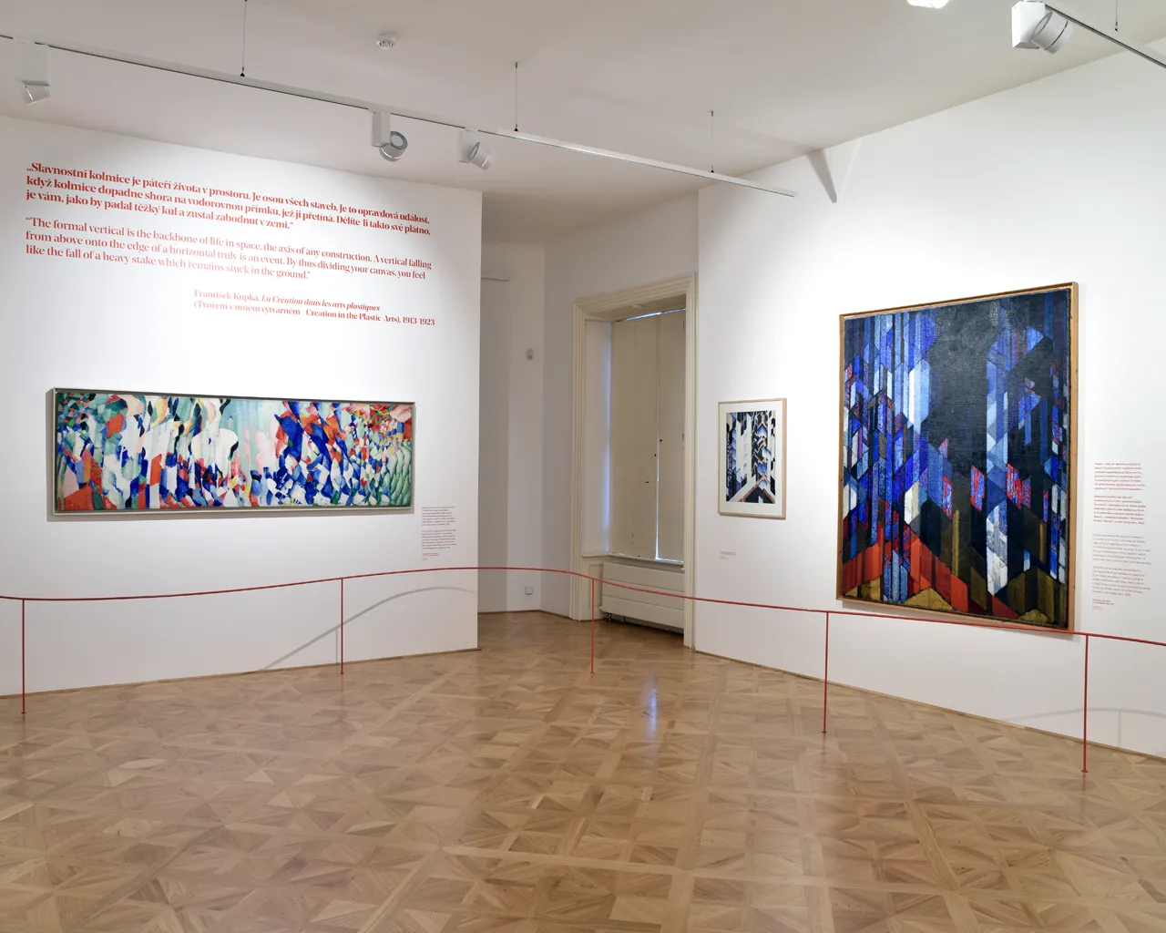 Una galería de arte con dos pinturas abstractas sobre paredes blancas, paneles de texto rojos y un suelo de madera