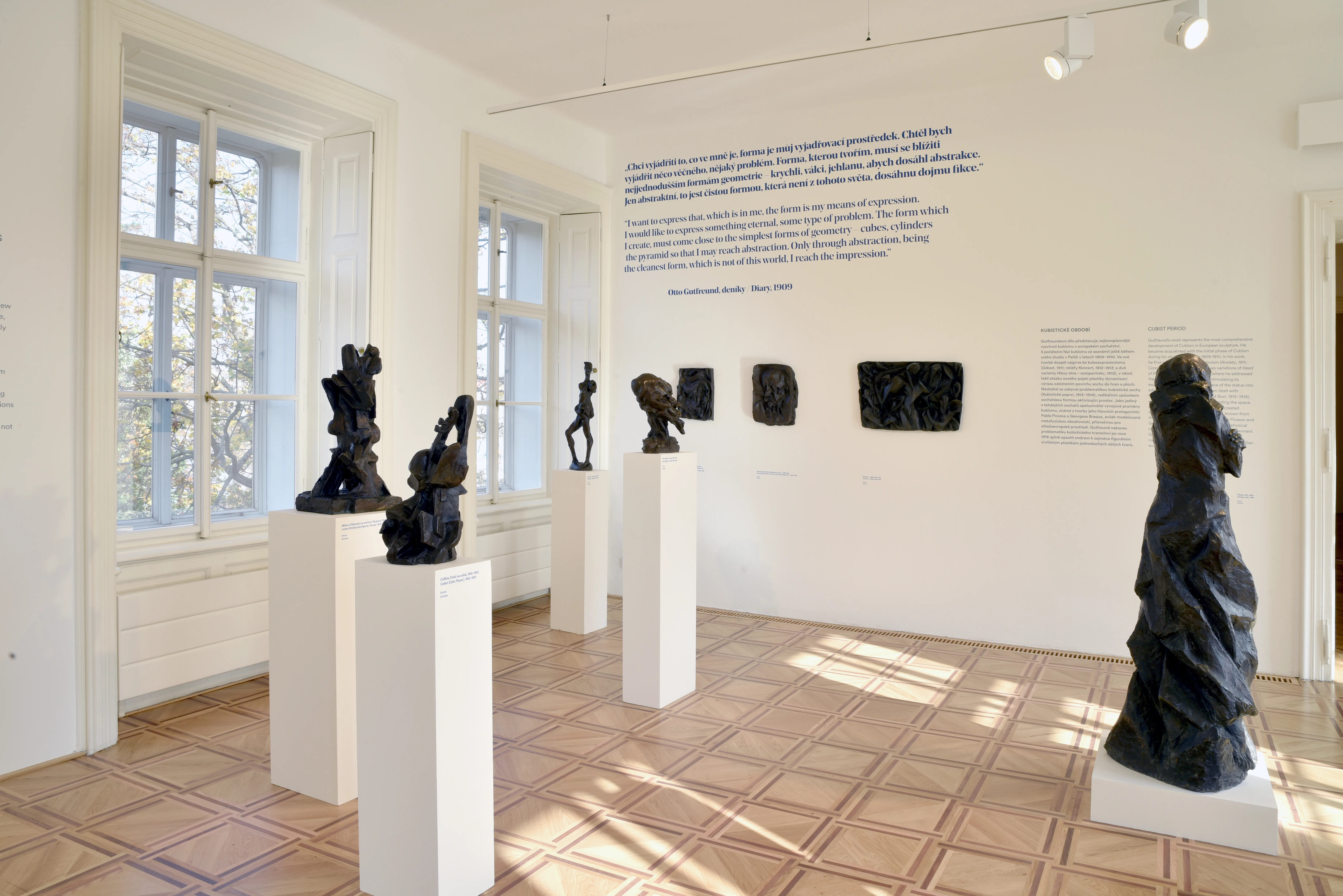 Esculturas sobre pedestales blancos en una galería con grandes ventanales y textos informativos en las paredes