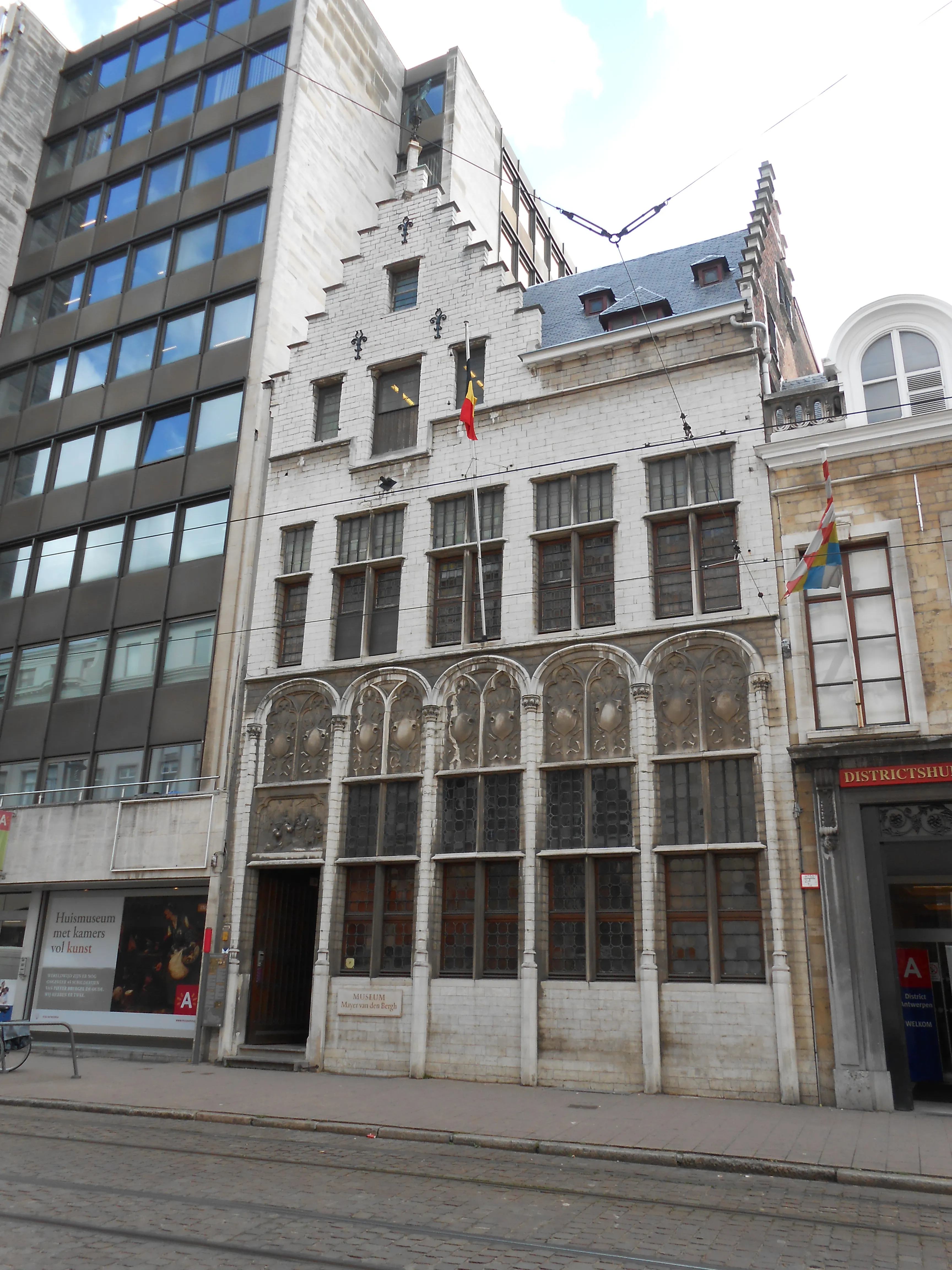 Edificio histórico de ladrillo blanco con fachada escalonada y ventanas arqueadas en la planta baja, que exhibe banderas de Bélgica y del arcoíris, situado entre edificios modernos y tradicionales en una calle con vías de tranvía.