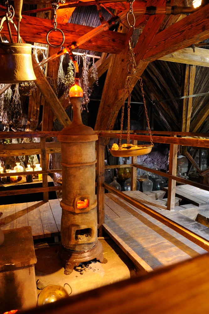 Interior of a historic alchemist's workshop with a central furnace, wooden beams, and hanging cauldrons