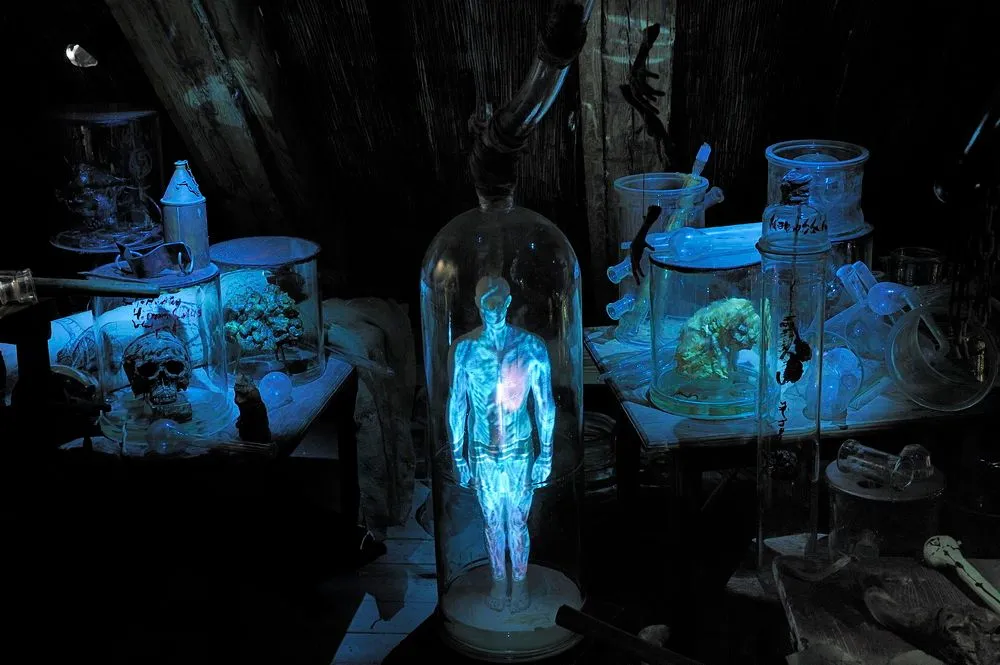 Glowing blue human figure in a glass dome surrounded by alchemical equipment and skulls