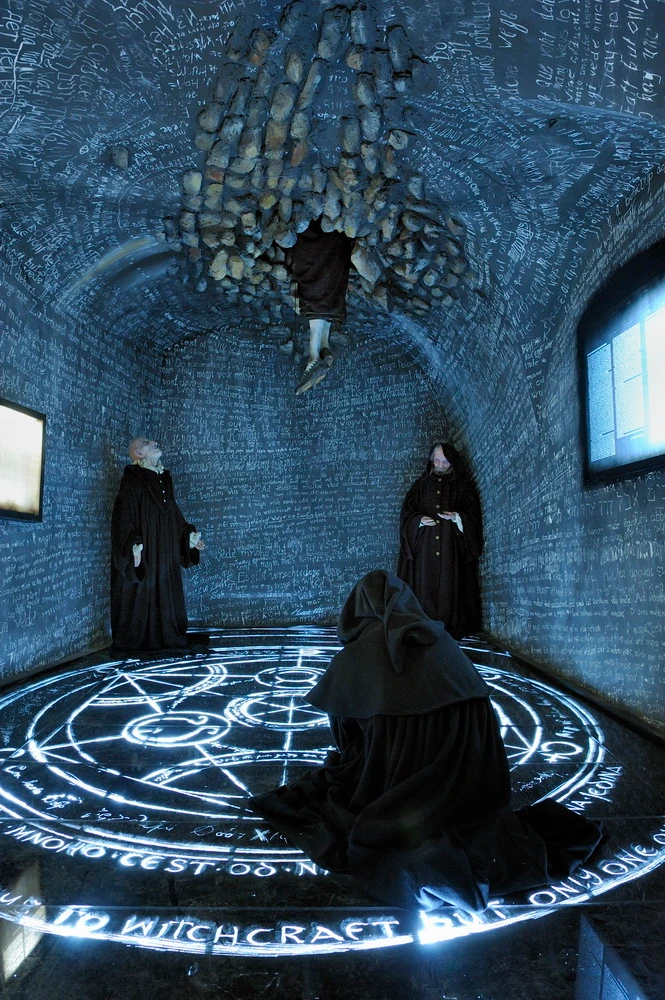 A dimly lit room with stone walls covered in script, three figures in black robes, and a glowing circular alchemical symbol on the floor.