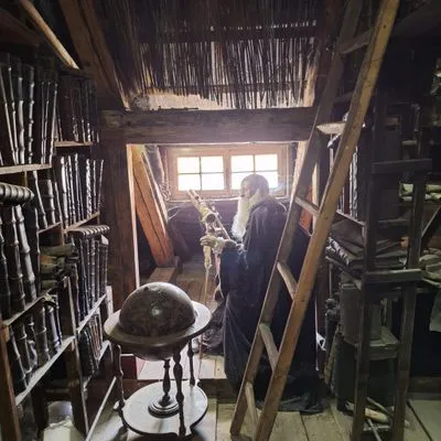 wooden bookshelves, a globe, a ladder, and a person in period clothing inside a dimly lit room