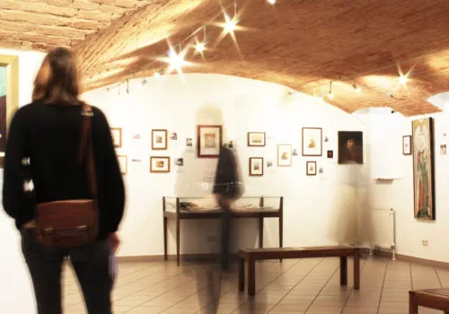 Person viewing framed artworks in a gallery with a wooden ceiling and display case