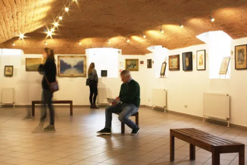 Interior of a museum gallery with people walking and sitting, framed artworks on white walls, wooden benches, and a vaulted ceiling with string lights.
