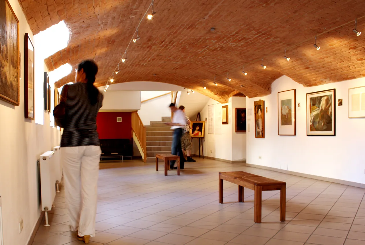Interior of a museum gallery with arched brick ceiling, white walls with framed artworks, wooden benches, and visitors walking