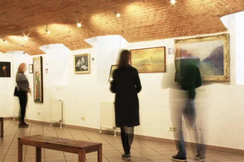 People viewing paintings in a gallery with white walls and a brick ceiling