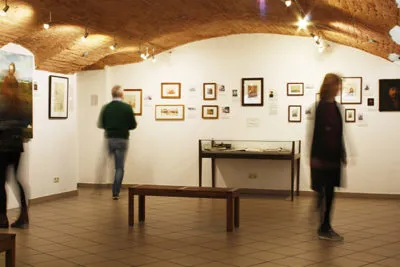 Interior of a museum gallery with visitors viewing framed artworks on white walls under arched ceilings.