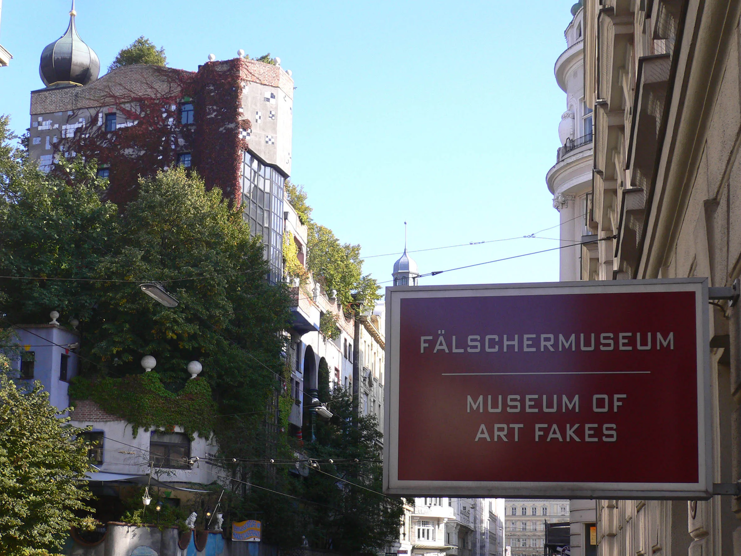 Red sign reading 'FÄLSCHERMUSEUM MUSEUM OF ART FAKES' on a building exterior with trees and other buildings in the background