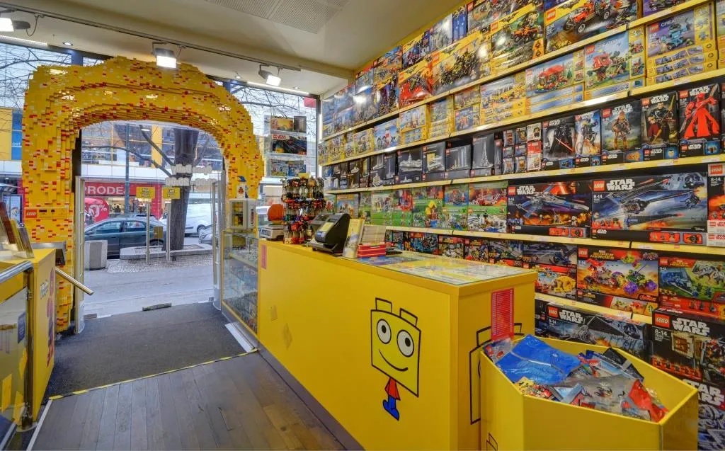 Gele receptiebalie met LEGO-thema en een lachend minifiguur-logo, omringd door schappen met LEGO-sets en een LEGO-boogvormige ingang