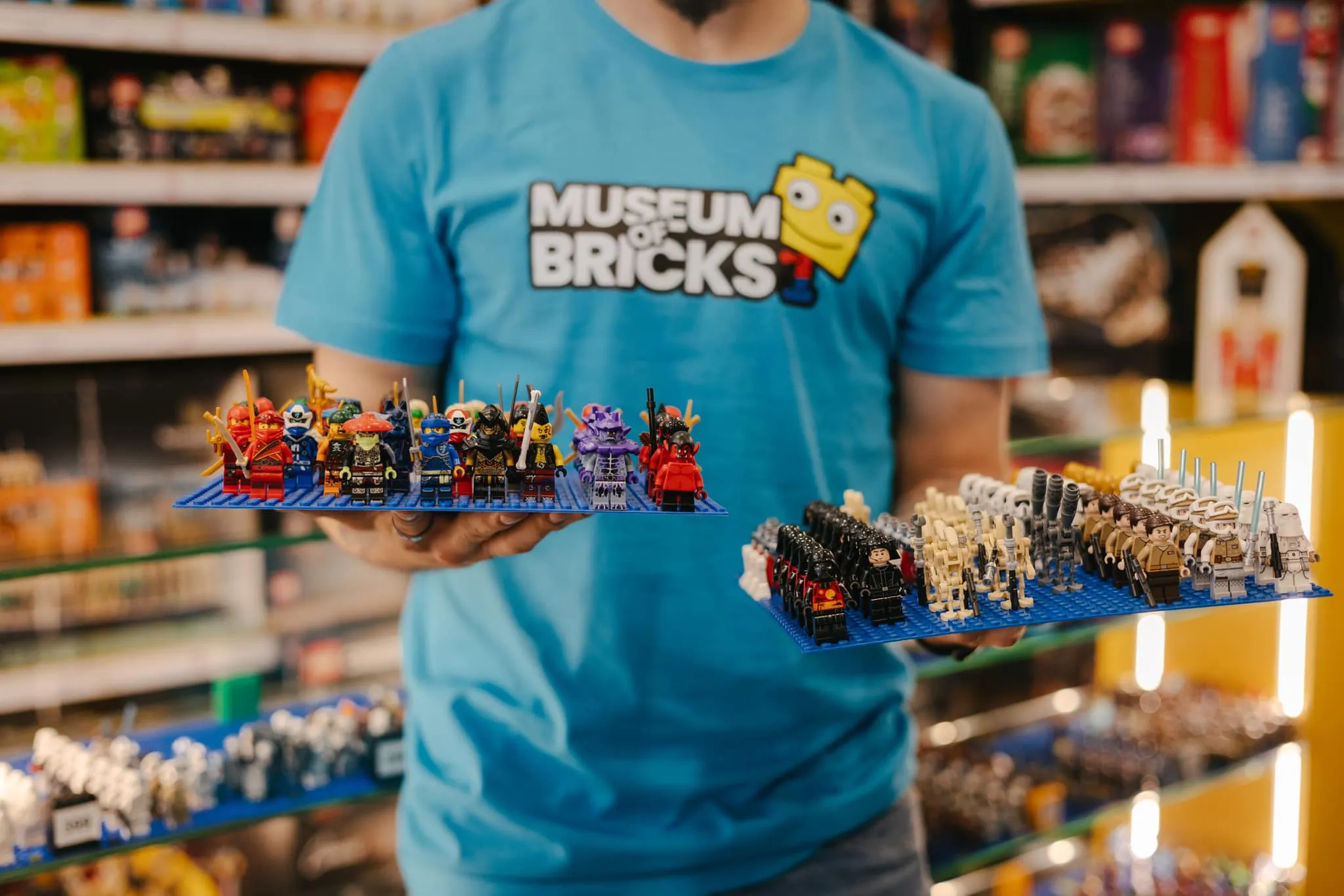 Een persoon in een blauw 'Museum of Bricks' T-shirt met twee bakjes LEGO-minifiguren in een museumsetting