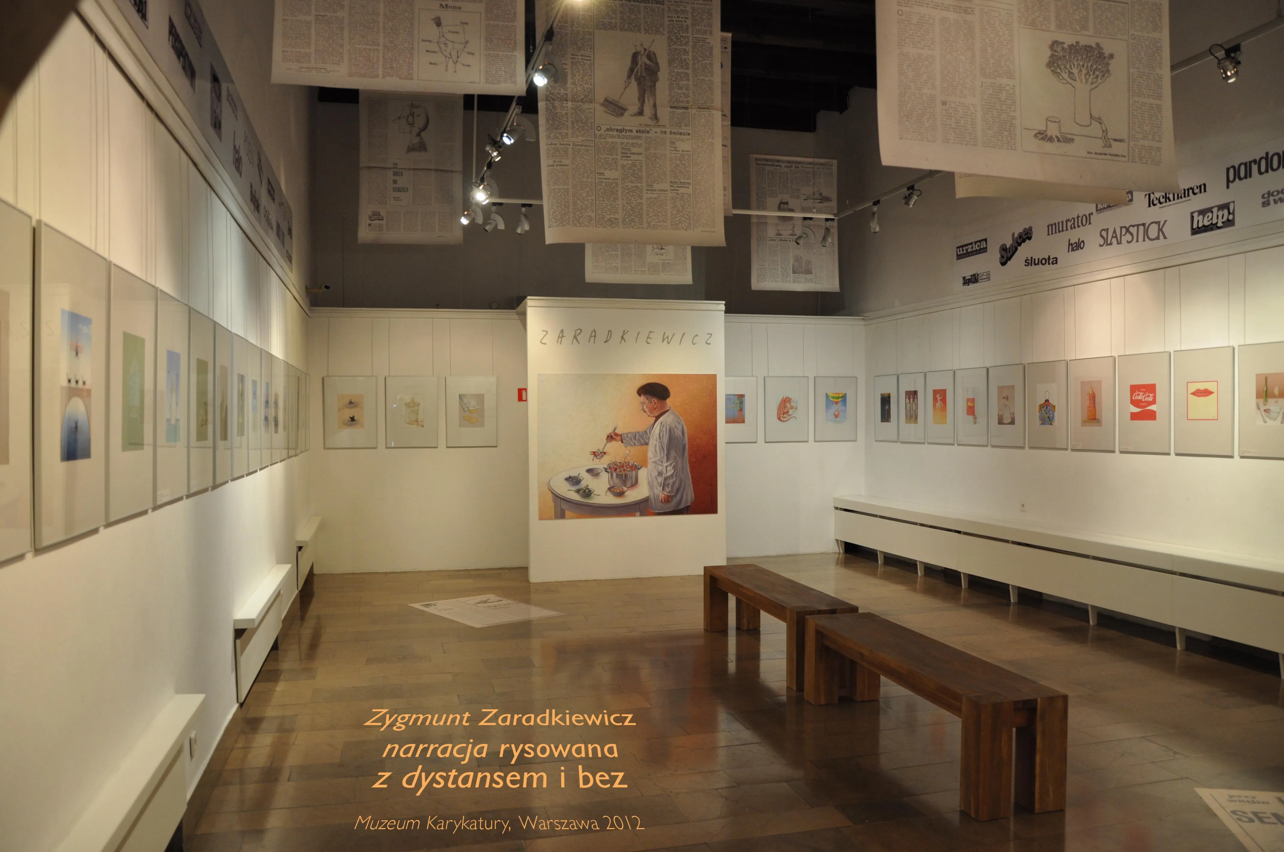 Espacio de exposición con obras de arte enmarcadas en paredes blancas, caricaturas grandes colgadas del techo y una exhibición central de una persona en una mesa.