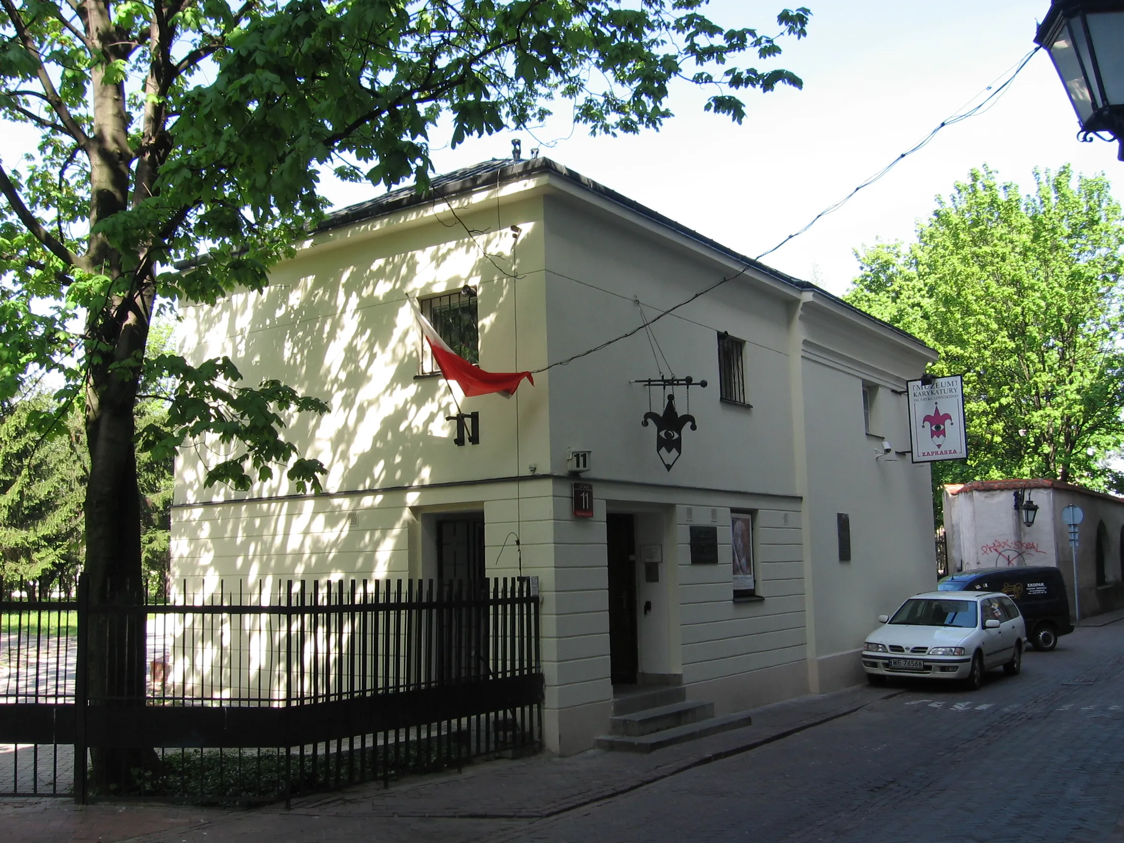 Un edificio de color claro con una bandera roja y un letrero decorativo, rodeado de árboles y una valla negra.