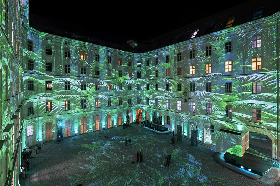 Proyecciones de luz verde con patrones de hojas en las paredes exteriores de un patio de edificio histórico por la noche