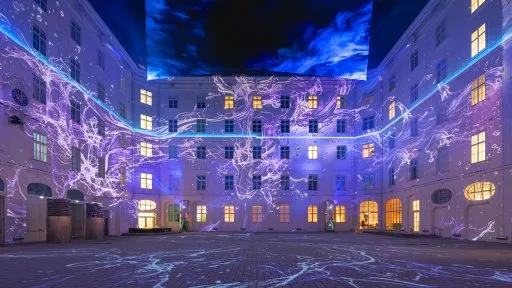 Un patio por la noche con un edificio grande que presenta ventanas iluminadas y proyecciones de luz morada y blanca en las paredes y el suelo.