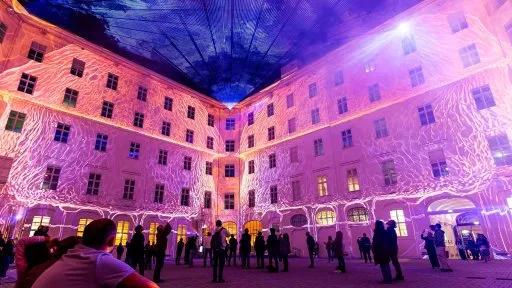 Un patio con un edificio grande iluminado por patrones de luz proyectada en púrpura y blanco, con gente reunida en primer plano.
