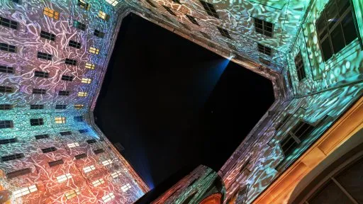 Proyecciones de luz colorida cubren las paredes interiores de un gran salón, creando una exhibición visual dinámica con patrones geométricos y un vacío central oscuro.