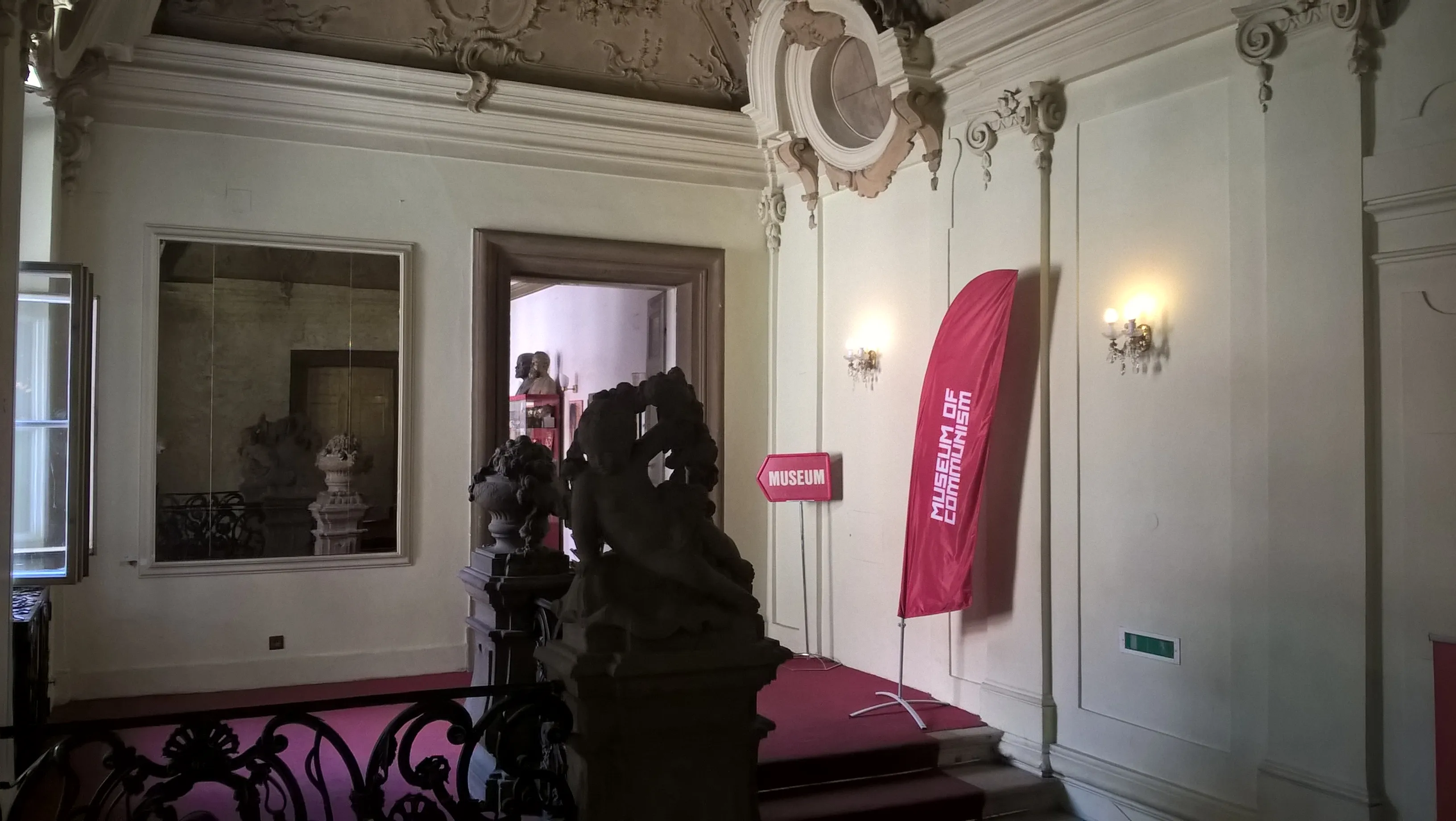 Interior ornamentado de un museo con paredes blancas, molduras decorativas, una pancarta roja y una escultura oscura en primer plano