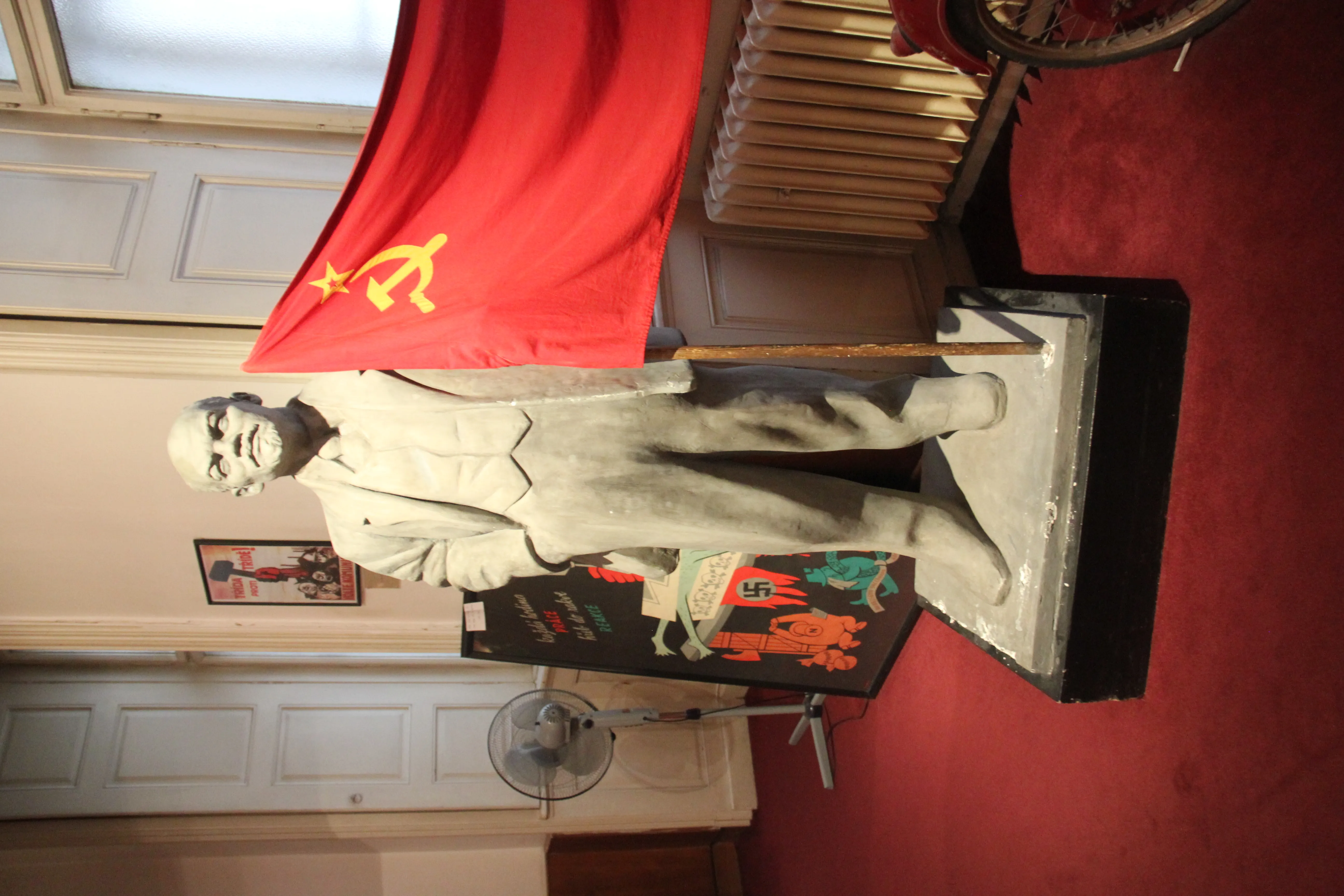 Estatua de un hombre con traje al lado de una bandera roja con un emblema de hoz y martillo, dentro de una exposición de museo