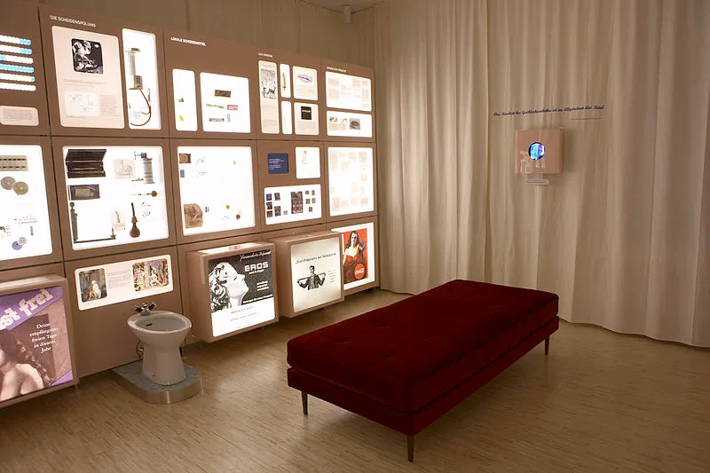 Sala de exposiciones con vitrinas iluminadas, un banco rojo y un dispositivo médico sobre un pedestal